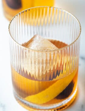 Old fashioned cocktail in a lowball glass, with single large cube of ice, cherry and orange peel in the glass.