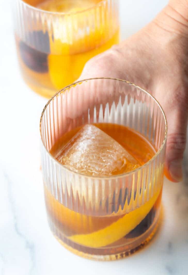 Hand holding an old fashioned cocktail in a lowball glass, with single large cube of ice, cherry and orange peel in the glass.