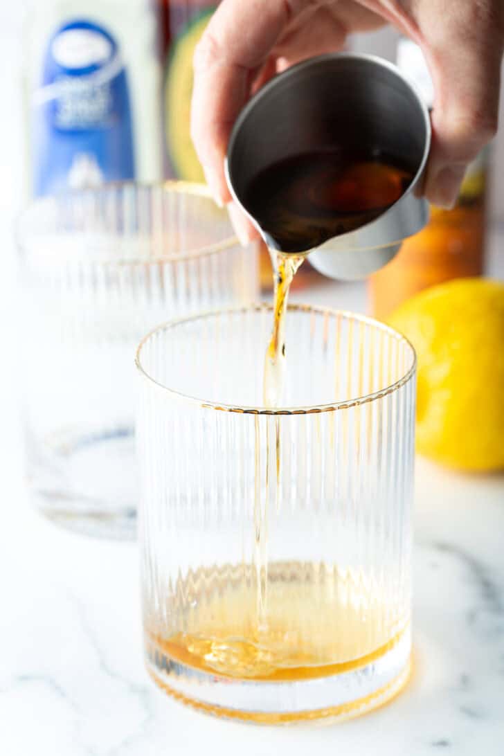 Pouring brown liquid from metal cup into a lowball glass.