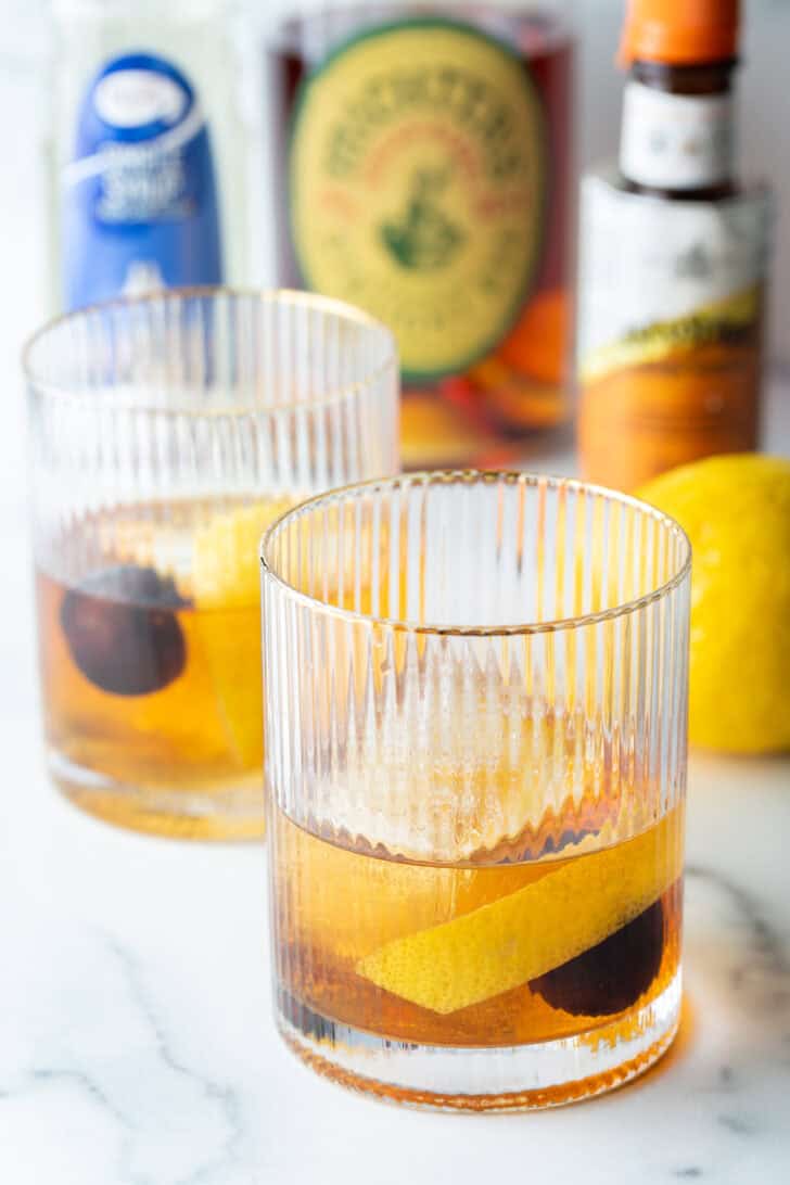 Old fashioned cocktail in a lowball glass, with single large cube of ice, cherry and orange peel in the glass.