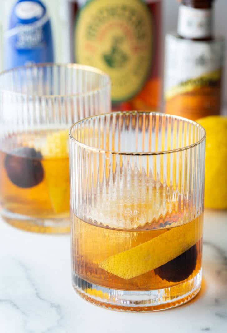 Old fashioned cocktail in a lowball glass, with single large cube of ice, cherry and orange peel in the glass.