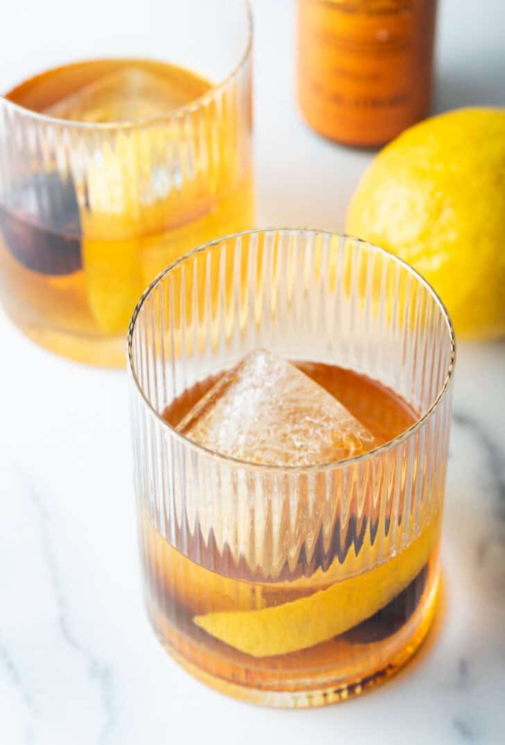 Old fashioned cocktail in a lowball glass, with single large cube of ice, cherry and orange peel in the glass.