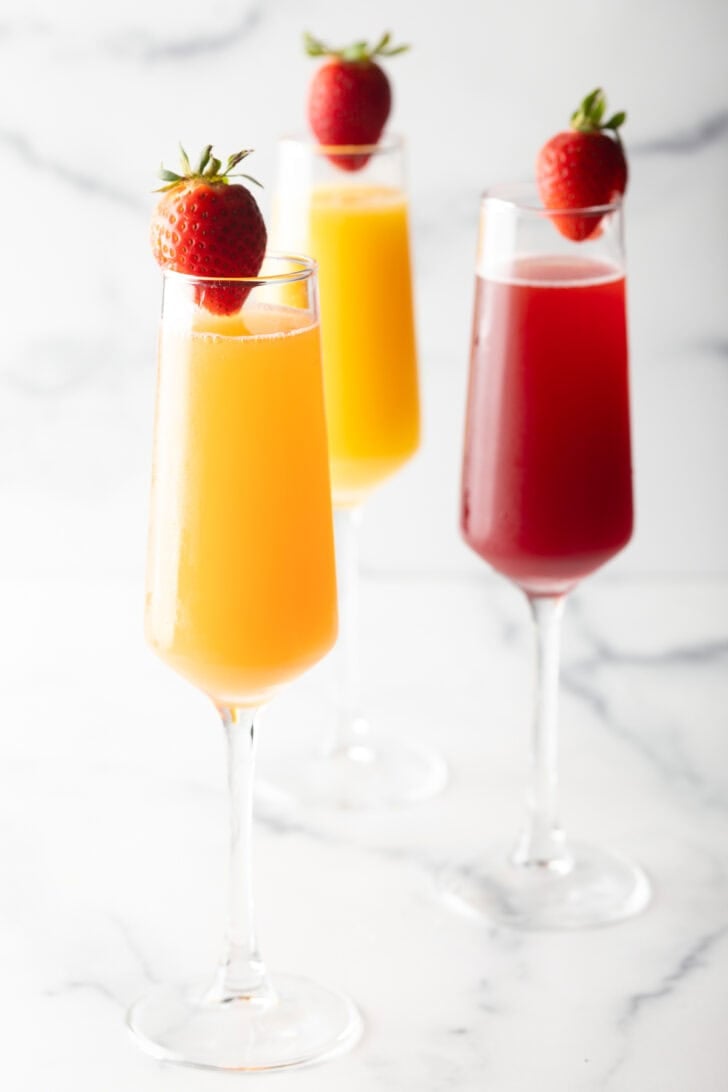 Two champagne flutes filled with orange mimosa, and another glass filled with a cranberry mimosa. Both glasses are garnished with a whole strawberry on the rim.