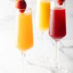 Two champagne flutes filled with orange mimosa, and another glass filled with a cranberry mimosa. Both glasses are garnished with a whole strawberry on the rim.