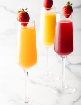 Two champagne flutes filled with orange mimosa, and another glass filled with a cranberry mimosa. Both glasses are garnished with a whole strawberry on the rim.