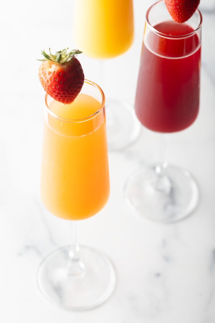 A champagne flute filled with orange mimosa, and another glass filled with a cranberry mimosa. Both glasses are garnished with a whole strawberry on the rim.