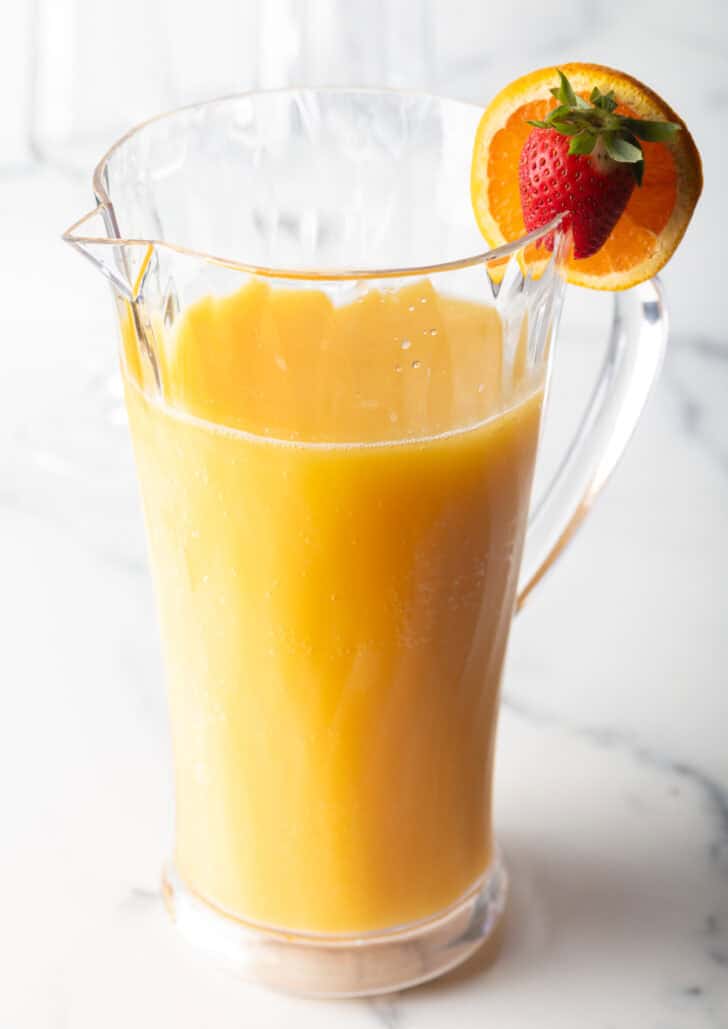 Large glass pitcher of mimosa with an orange and strawberry garnish on the rim.
