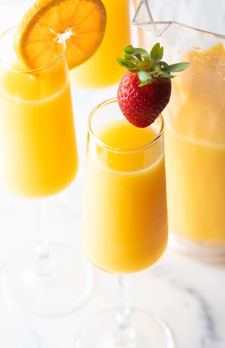 Two glasses of mimosas, one garnished with a large strawberry on the rim, the other garnished with an orange slice on the rim.
