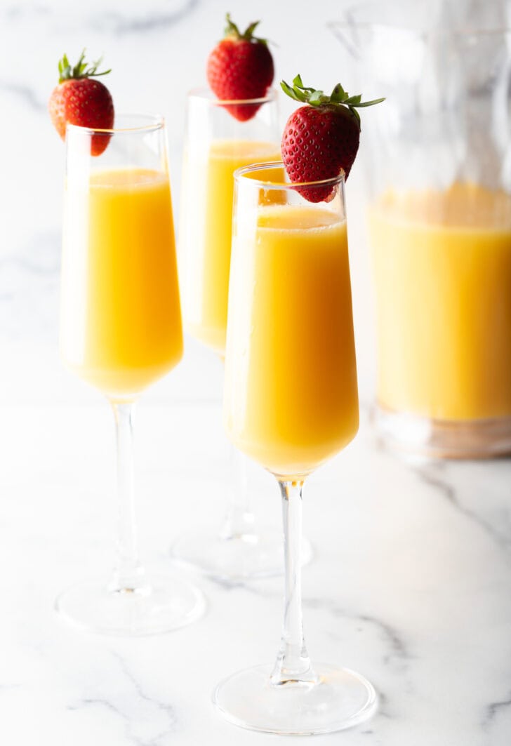 3 champagne glasses full of mimosa and each garnished with a whole strawberry on the rim.