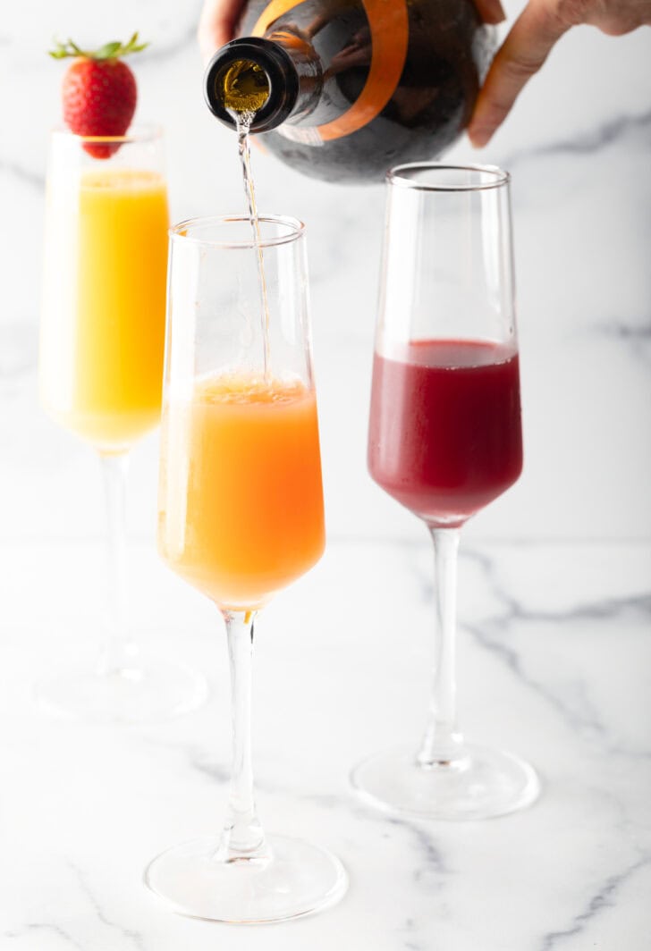 3 champagne glass against a white marble backdrop. The glass in the background is full of orange juice and garnished with a whole strawberry on the rim. In the foreground are a glass half full with cranberry juice, and a glass half full of orange juice with sparkling wine being poured in to the glass.
