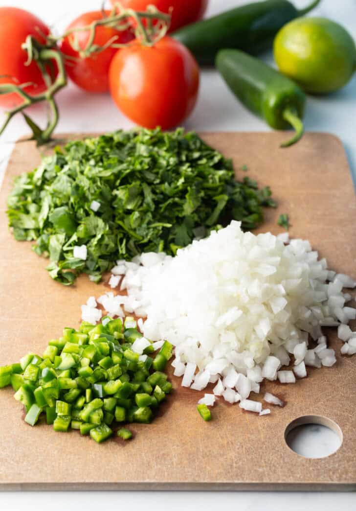 Recipe ingredients in separate piles on a cutting board: Fresh chopped cilantro, jalapenos, and onion.