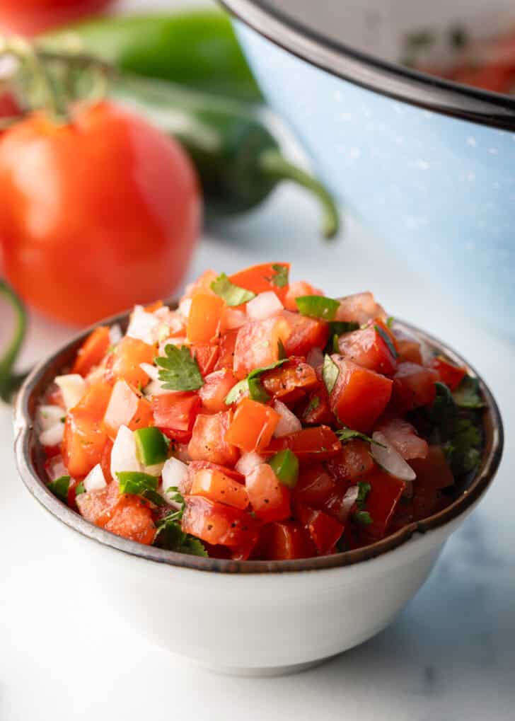 A white bowl of fresh chopped tomatoes, onion, jalapeno, and cilantro. The bowl is surrounded by tortilla chips.