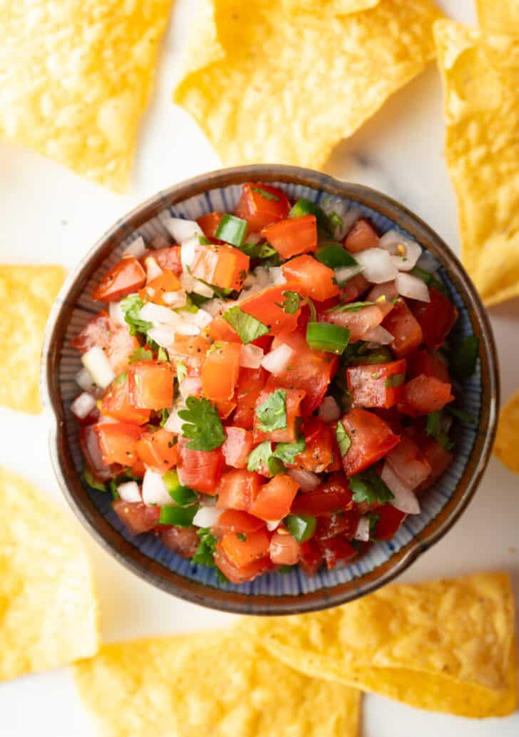 Top down view of a white bowl of fresh pico de gallo with chopped tomatoes, onion, jalapeno, and cilantro. The bowl is surrounded by tortilla chips.