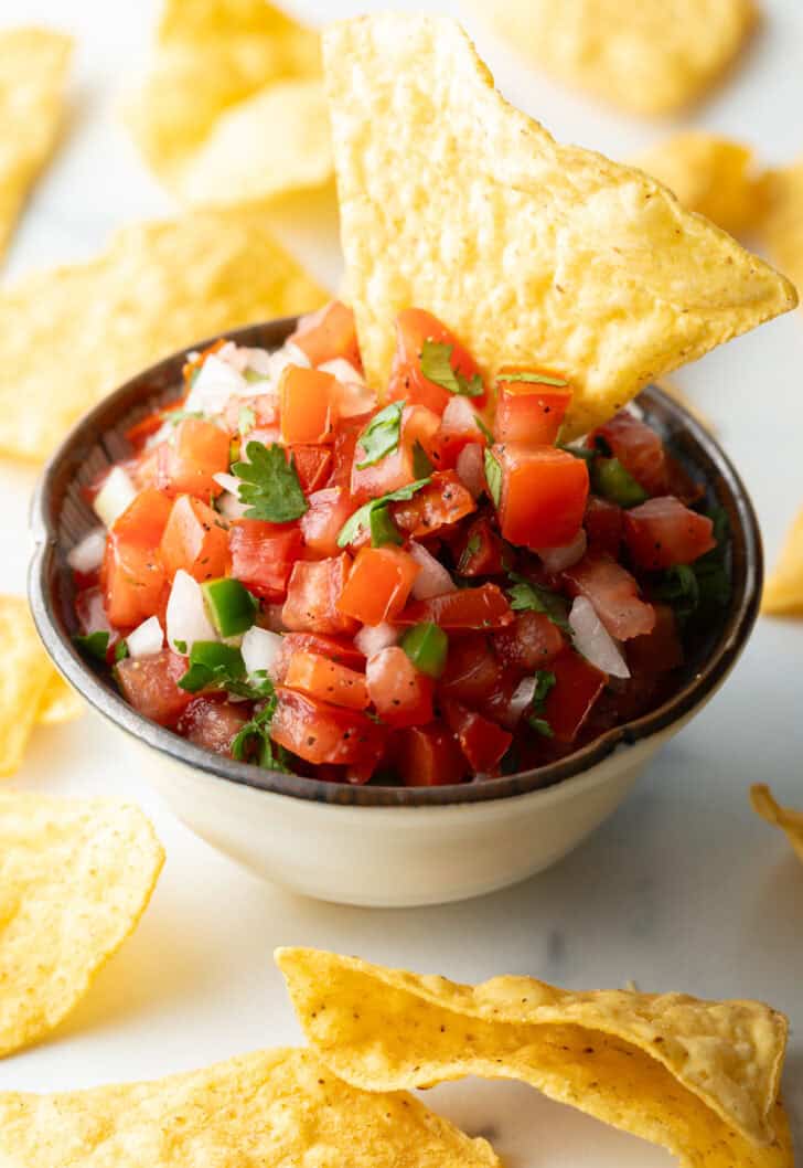 A white bowl of fresh pico de gallo with chopped tomatoes, onion, jalapeno, and cilantro. A tortilla chip is sticking out of the bowl.