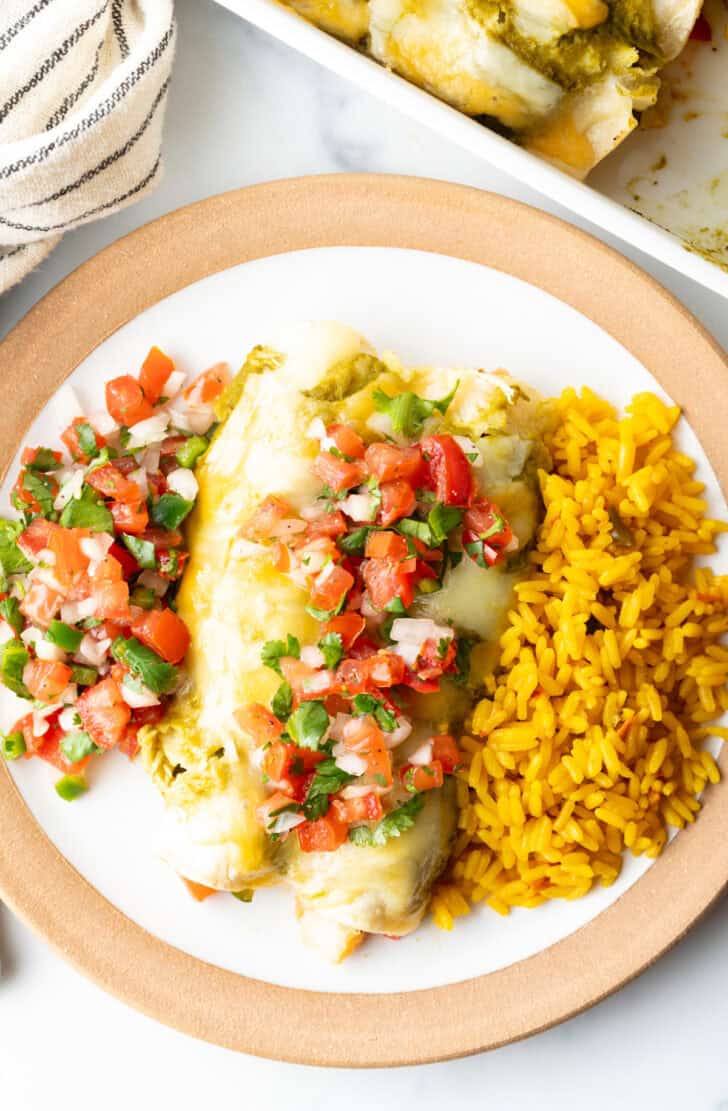 Top down view of two enchiladas next to a serving of yellow rice. A scoop of pico de gallo is on the other side of the enchiladas, and a scoop is also on top of the enchiladas.