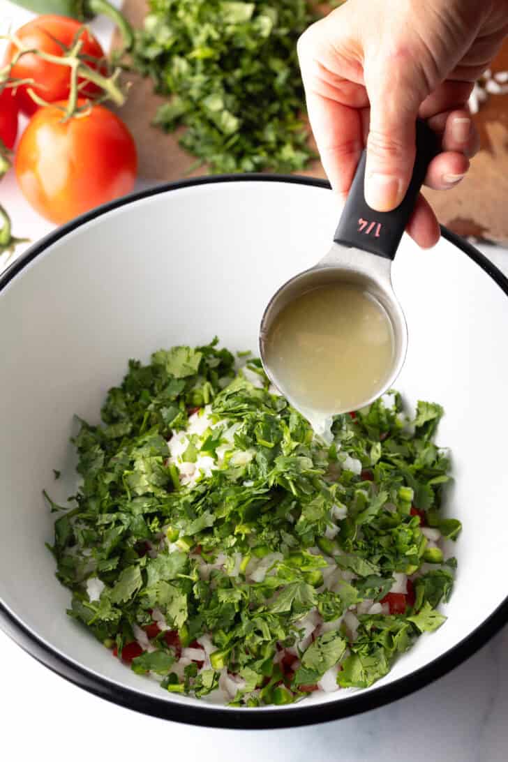 Fresh chopped cilantro has been added to the bowl. A hand is pouring lime juice from a small metal measuring cup.