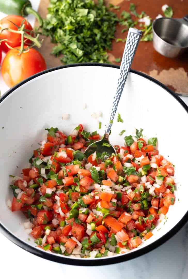 Combining the pico do gallo ingredients in a white mixing bowl with a spoon.