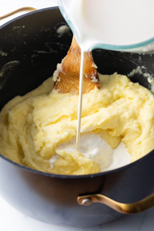 Adding heavy cream to a large pot of cooked potatoes.