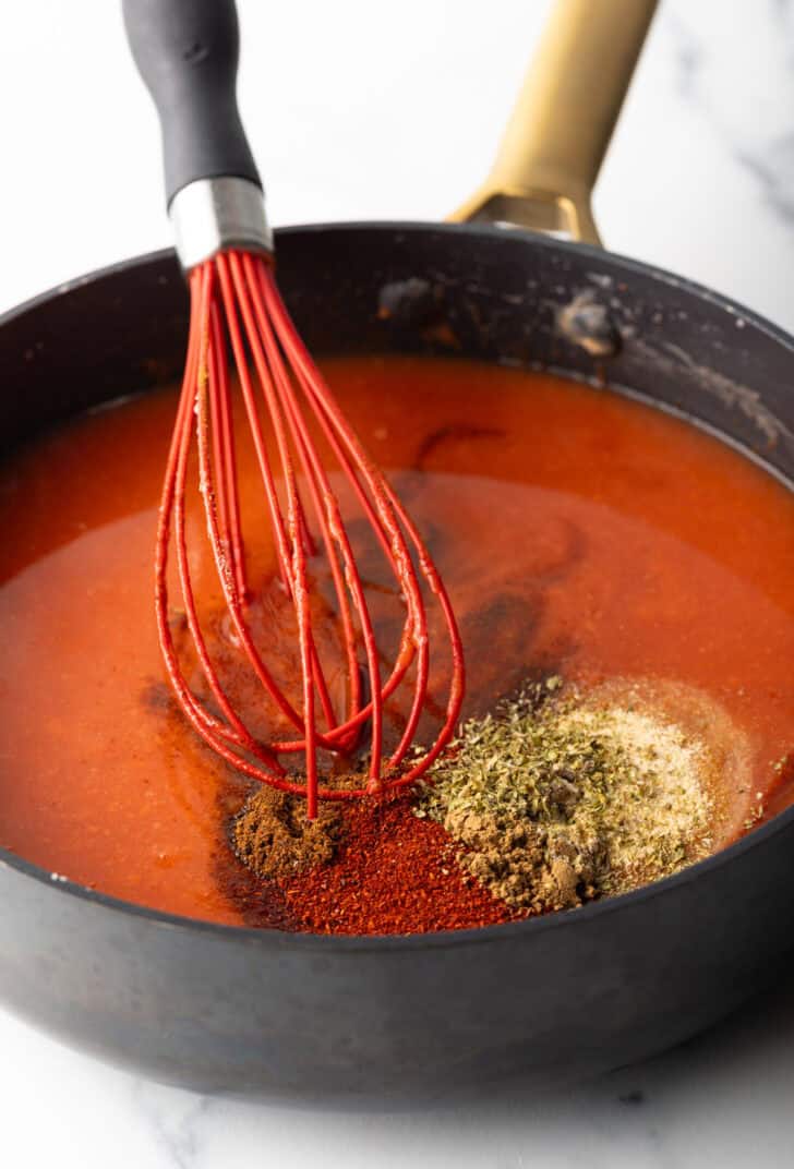 Large deep skillet with red sauce, spices, and a red whisk.