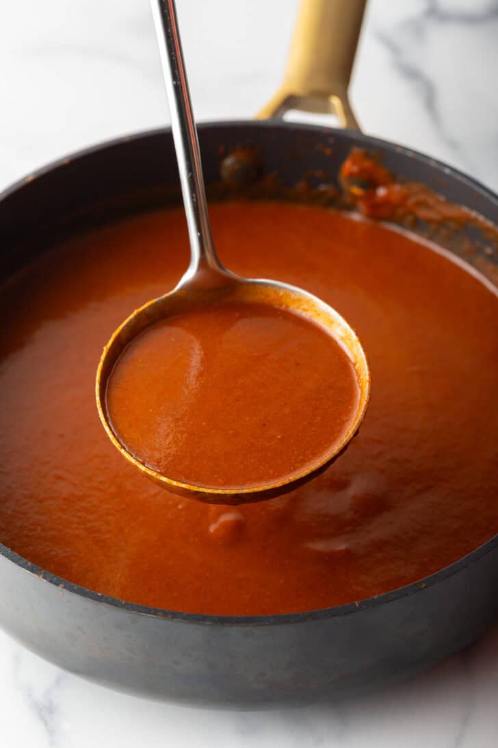 Metal ladleful of red sauce being held above a black pan of sauce.