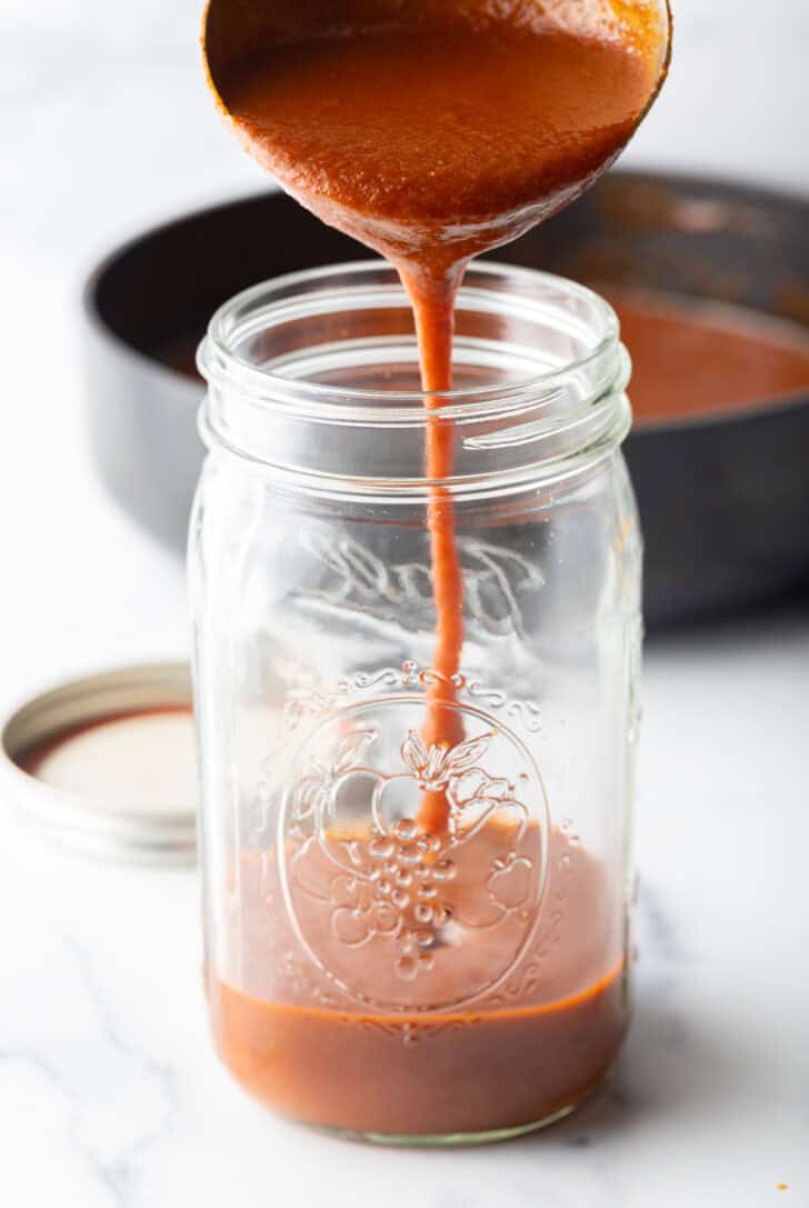 Pouring red sauce from a ladle into a glass mason jar.