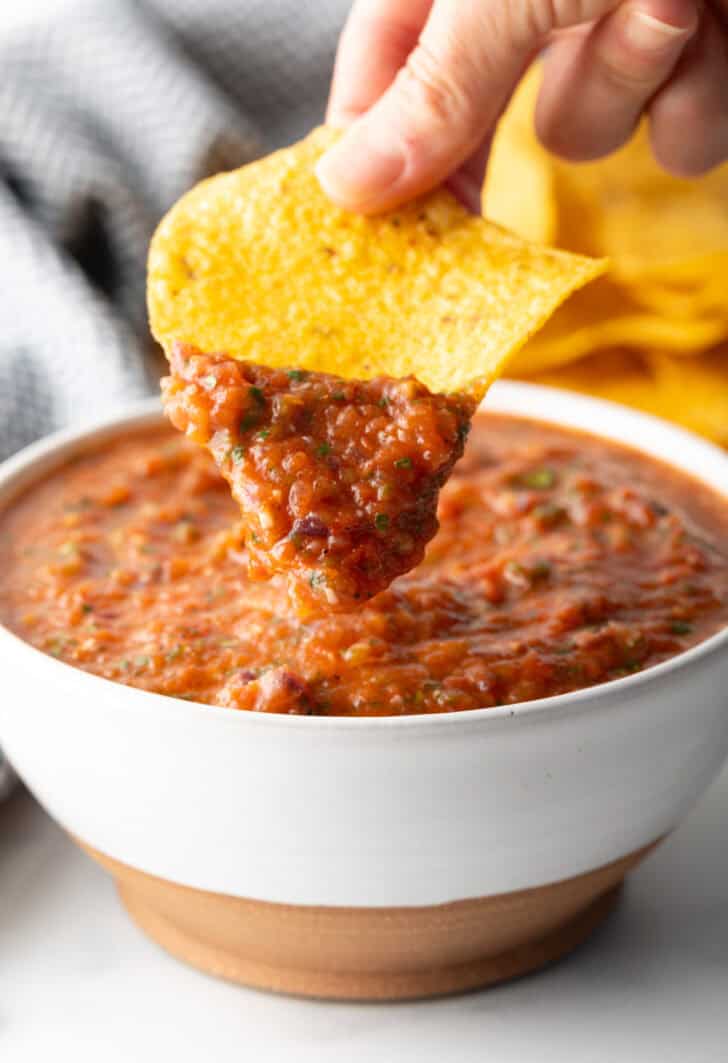 White bowl of roasted tomato salsa. A hand is showing a scoop of salsa on a tortilla chip to camera.
