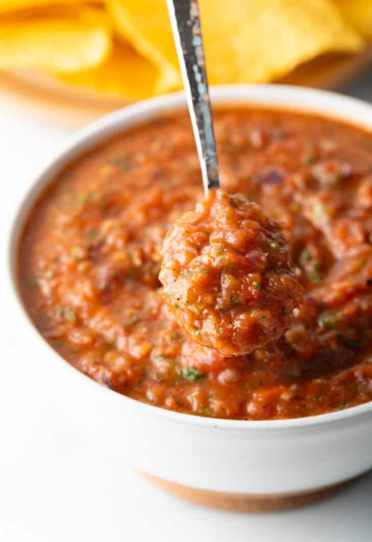 A metal spoon is showing a scoop of roasted salsa scooped from a white bowl.