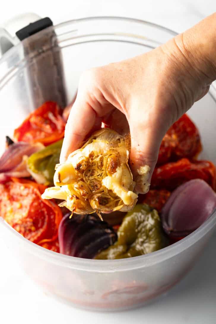 Hand squeezing roasted garlic into a food processor with other salsa ingredients.
