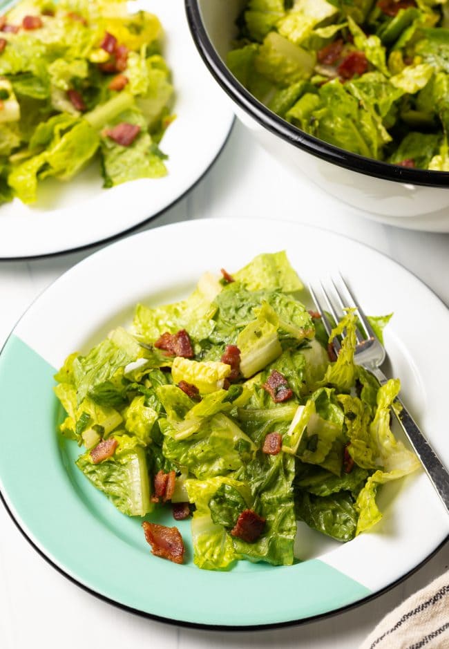 top side view of wilted lettuce salad with chopped bacon on a white and teal plate and fork on the side