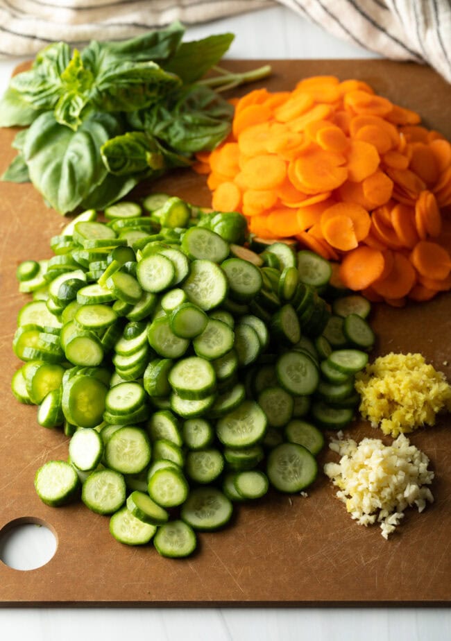 Recipe ingredients: Carrots, mini cucumbers, fresh ginger. All ingredients are sliced and chopped on a cutting board.