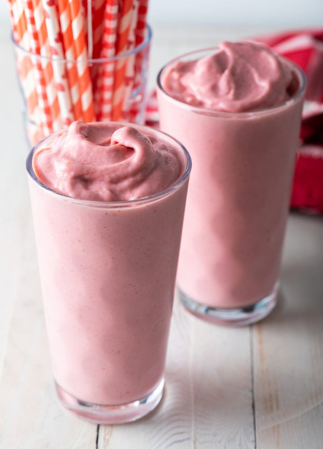 Two glasses of strawberry smoothie with a jar of straws in the background.