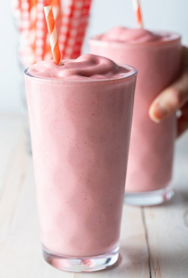 Two strawberry smoothies with a hand grabbing one of the glasses. 