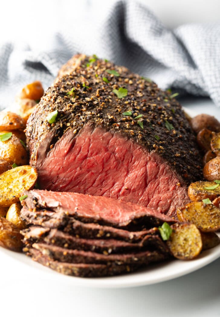 Cooked beef round roast on a metal baking pan. The front half of the roast has been thinly sliced and the meat is surrounded by roasted baby potatoes.