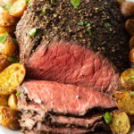 Cooked beef round roast on a metal baking pan. The front half of the roast has been thinly sliced and the meat is surrounded by roasted baby potatoes.