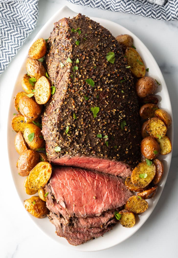 Top down view cooked beef round roast on a metal baking pan. The front half of the roast has been thinly sliced and the meat is surrounded by roasted baby potatoes.
