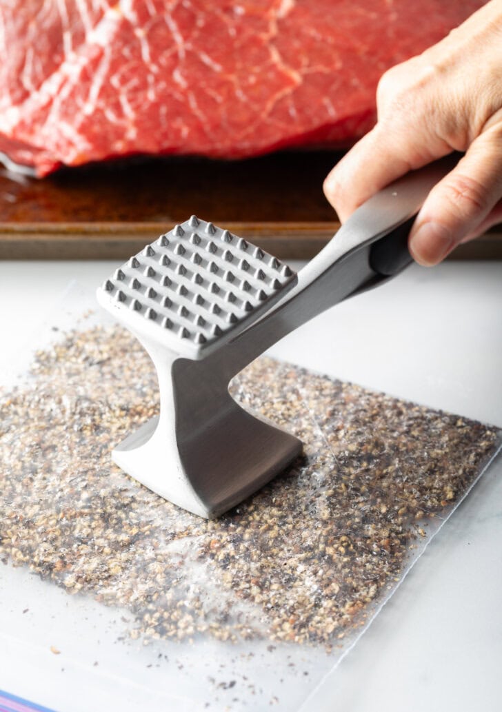 Hand with metal mallet smashed peppercorns in a plastic zipper bag.