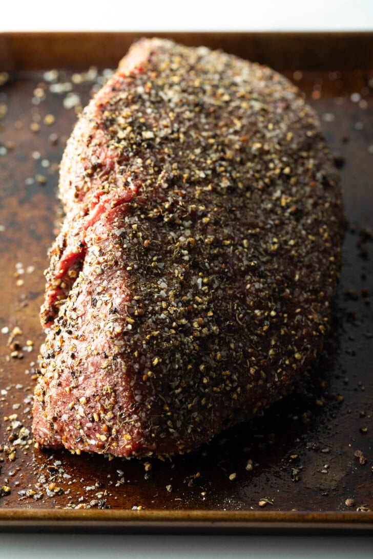Raw top round roast coated with pepper rub, on a baking sheet.