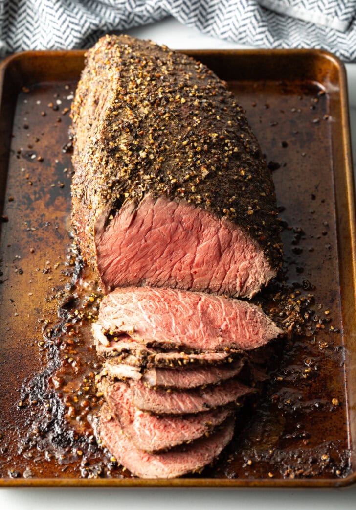 Top down view cooked beef top round roast on a metal baking pan. The front half of the roast has been thinly sliced.