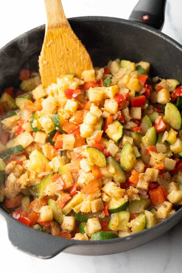 Diced sauteed peppers, zucchini, potatoes and onions being stirred with a wood spatula.