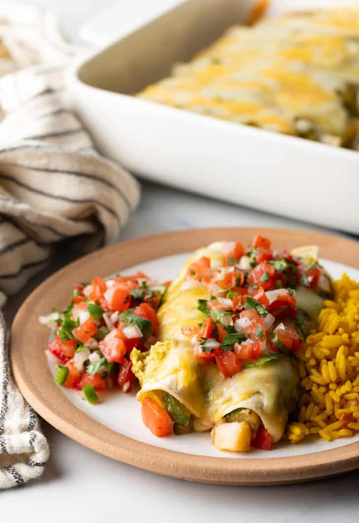 White plate with a serving of veggie enchiladas with green sauce and pico de gallo, nestled between a serving of pico de gallo and spanish rice.