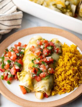 White plate with a serving of veggie enchiladas with green sauce and pico de gallo, nestled between a serving of pico de gallo and spanish rice.