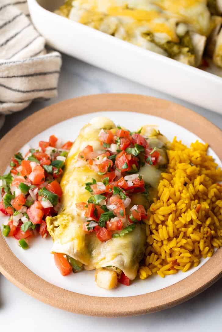 White plate with a serving of veggie enchiladas with green sauce and pico de gallo, nestled between a serving of pico de gallo and spanish rice.
