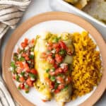 Top down view white plate with a serving of veggie enchiladas with green sauce and pico de gallo, nestled between a serving of pico de gallo and spanish rice.