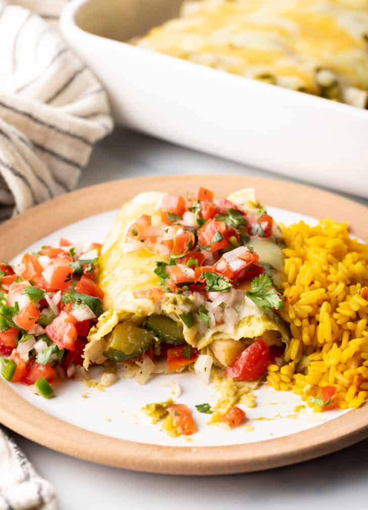 White plate with a serving of veggie enchiladas with green sauce and pico de gallo, nestled between a serving of pico de gallo and spanish rice.