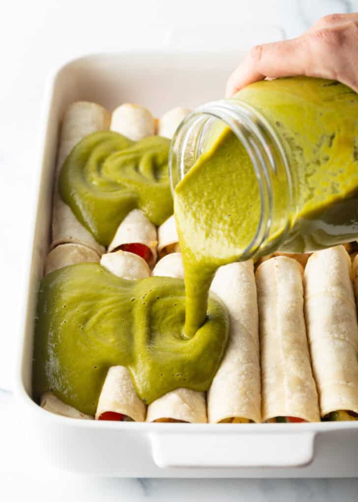 Pouring green sauce from a large glass over two rows of enchiladas in a white baking dish.