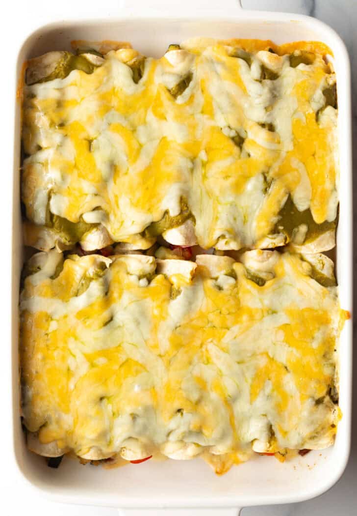Top down view of a dozen vegetable enchiladas covered in green sauce and cheese in a white baking dish.