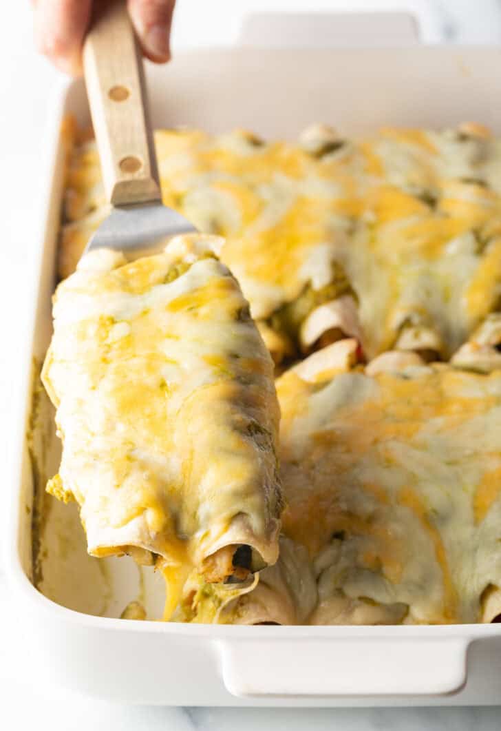 Hand with a metal spatula removing two enchiladas from a baking dish.