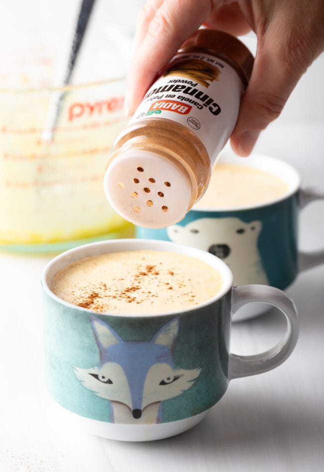 Adding a sprinkle of cinnamon to a mug of frothy coffee.